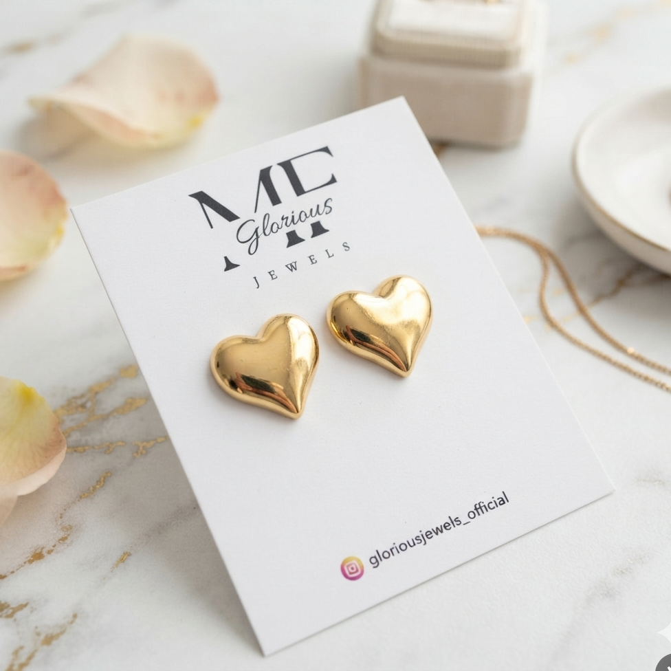 A pair of shiny gold heart-shaped stud earrings displayed on a white jewelry card labeled 'ME Glorious Jewels', set on a light marble surface with soft rose petals and jewelry props around it.