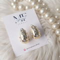 A pair of gold teardrop-shaped earrings with rhinestone embellishments, displayed on a white card labeled 'MC Glorious Jewels' on a white faux fur surface with pearl strands in the background.