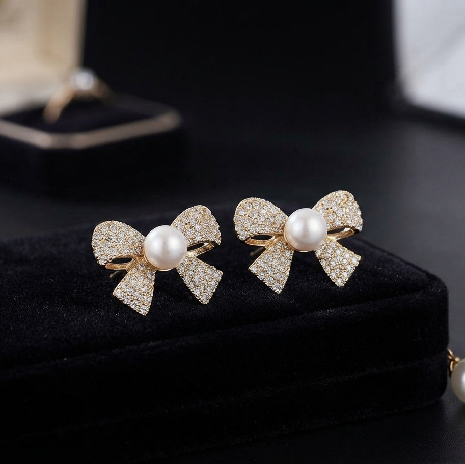 A pair of gold bow-shaped earrings with pavé-set clear stones and a central white pearl, displayed on a black velvet jewelry box.