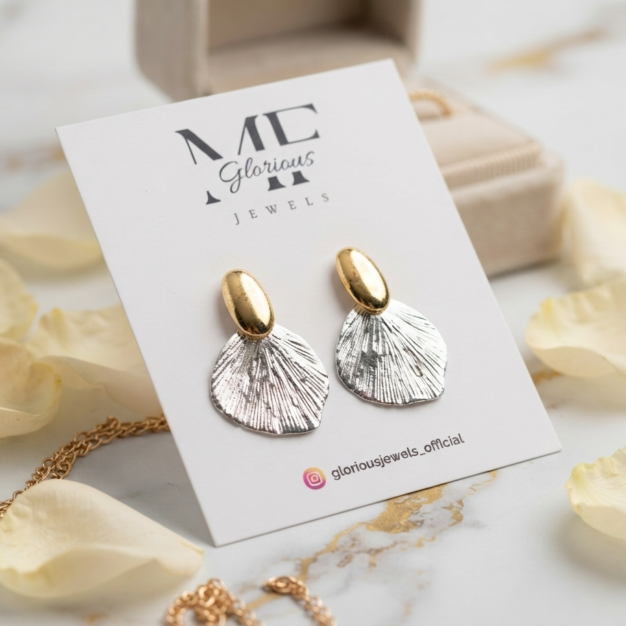 A pair of gold and silver shell-shaped drop earrings displayed on a white branded card, styled with cream rose petals and a gold necklace on a light marble surface.