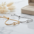 Two adjustable chain bracelets with evil eye motifs, one in yellow gold and one in silver, displayed on a white marble surface with a beige jewelry box and dried flowers in the background.