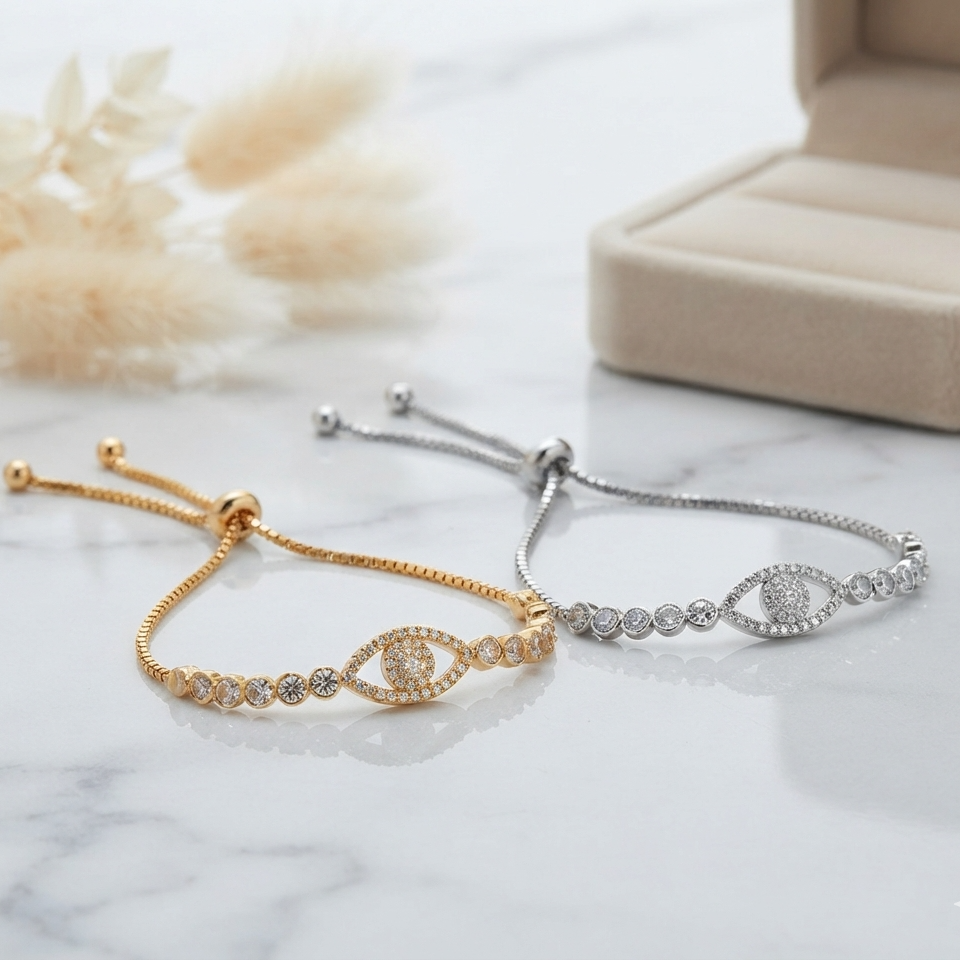 Two adjustable chain bracelets with evil eye motifs, one in yellow gold and one in silver, displayed on a white marble surface with a beige jewelry box and dried flowers in the background.