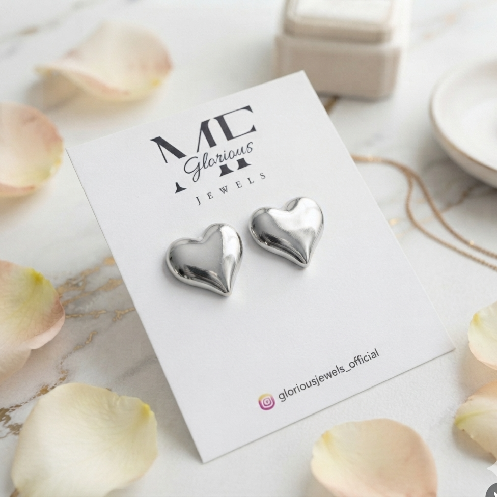 A pair of shiny silver heart-shaped stud earrings displayed on a white jewelry card labeled 'ME Glorious Jewels,' styled on a light marble surface with soft rose petals and a ring box in the background.