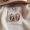 A pair of double-loop hoop earrings in gold and silver tones, displayed on a white jewelry card labeled 'ME Glaciers Jewelry', styled on a white fabric surface with an open book and pearl necklace in the background.
