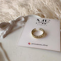 A gold-colored textured ring displayed on a white branded card, set on a white surface with a white bow and fluffy white fabric in the background.