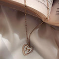 A rose gold chain necklace with a heart-shaped pendant featuring a Roman numeral 'I' surrounded by small clear rhinestones, draped over an open book on a white satin fabric background.