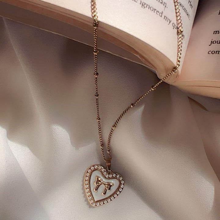 A rose gold chain necklace with a heart-shaped pendant featuring a Roman numeral 'I' surrounded by small clear rhinestones, draped over an open book on a white satin fabric background.