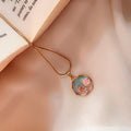 A gold necklace with a round pendant featuring a pastel illustration of a pink flower on a light blue background, resting on a soft beige fabric next to an open book.