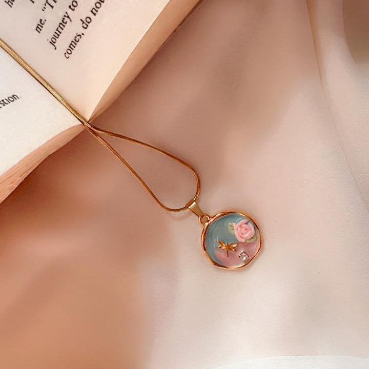 A gold necklace with a round pendant featuring a pastel illustration of a pink flower on a light blue background, resting on a soft beige fabric next to an open book.