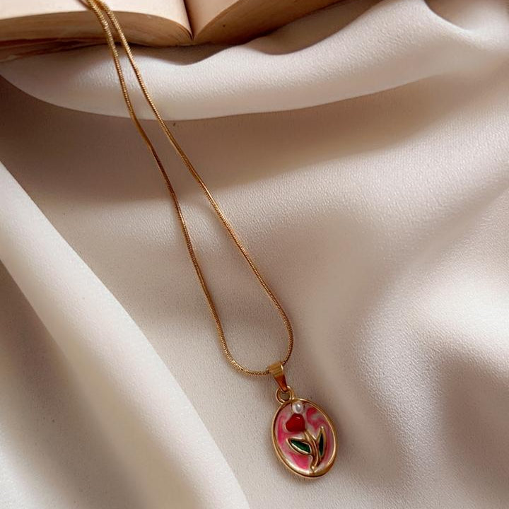 A gold chain necklace with an oval pendant featuring a red rose with green leaves, styled on draped white fabric next to an open book.