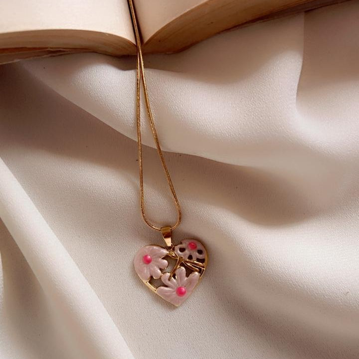 A gold heart-shaped pendant necklace with pink glitter and a small cross, draped over a cream satin fabric next to an open book.