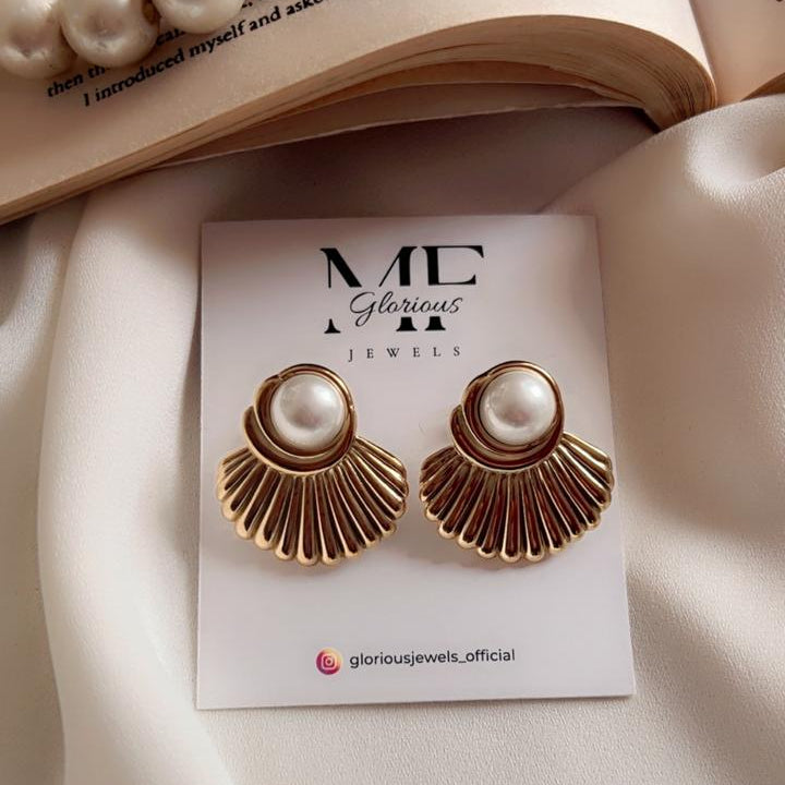 A pair of gold-toned fan-shaped earrings with round white pearl centers, displayed on a white jewelry card. The card is placed on draped white fabric with an open book and pearl beads in the background.
