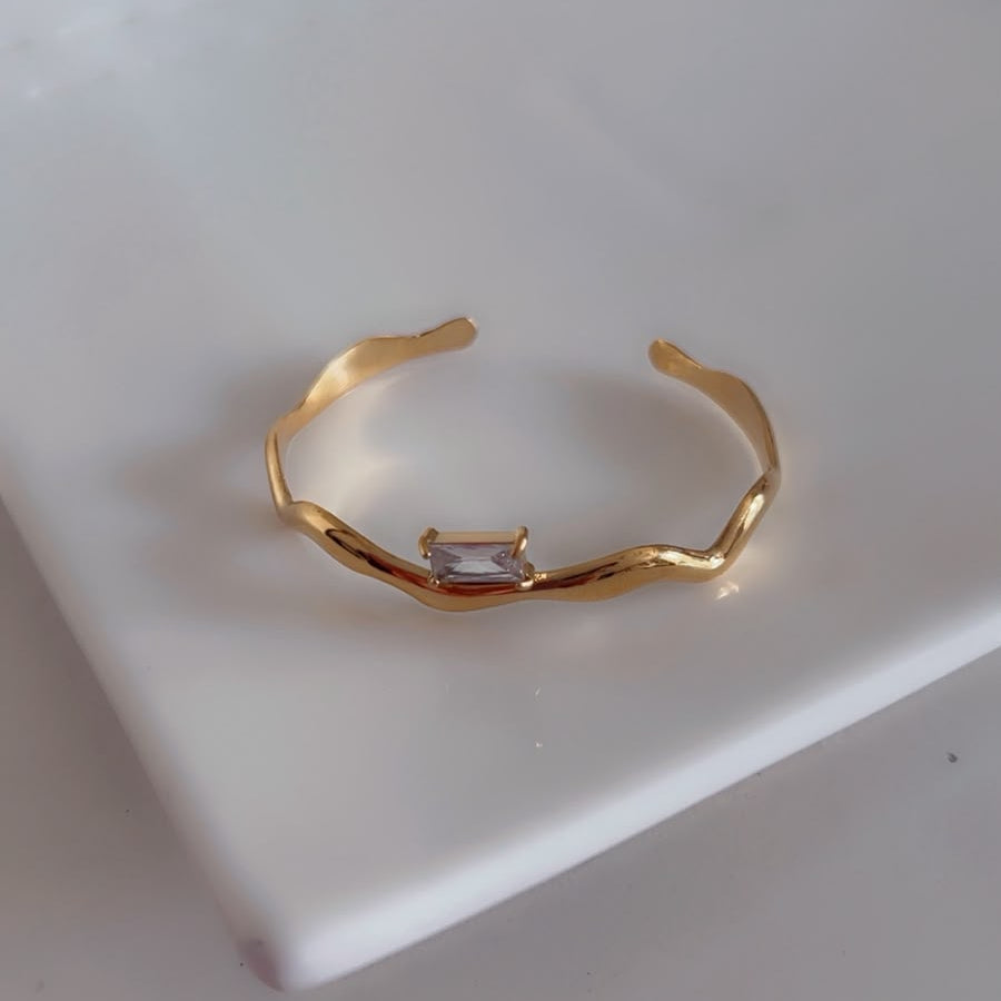 A gold open cuff bracelet with a wavy, organic design and a small rectangular clear gemstone in the center, resting on a white ceramic dish.