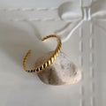 A gold twisted wire cuff bracelet resting on a small light-colored stone, placed on a white textured surface with a bow design in the background.