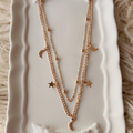 Two delicate gold-tone layered necklaces with crescent moon and star charms, displayed on a white ceramic tray with bow details, set against a fluffy cream faux fur background.